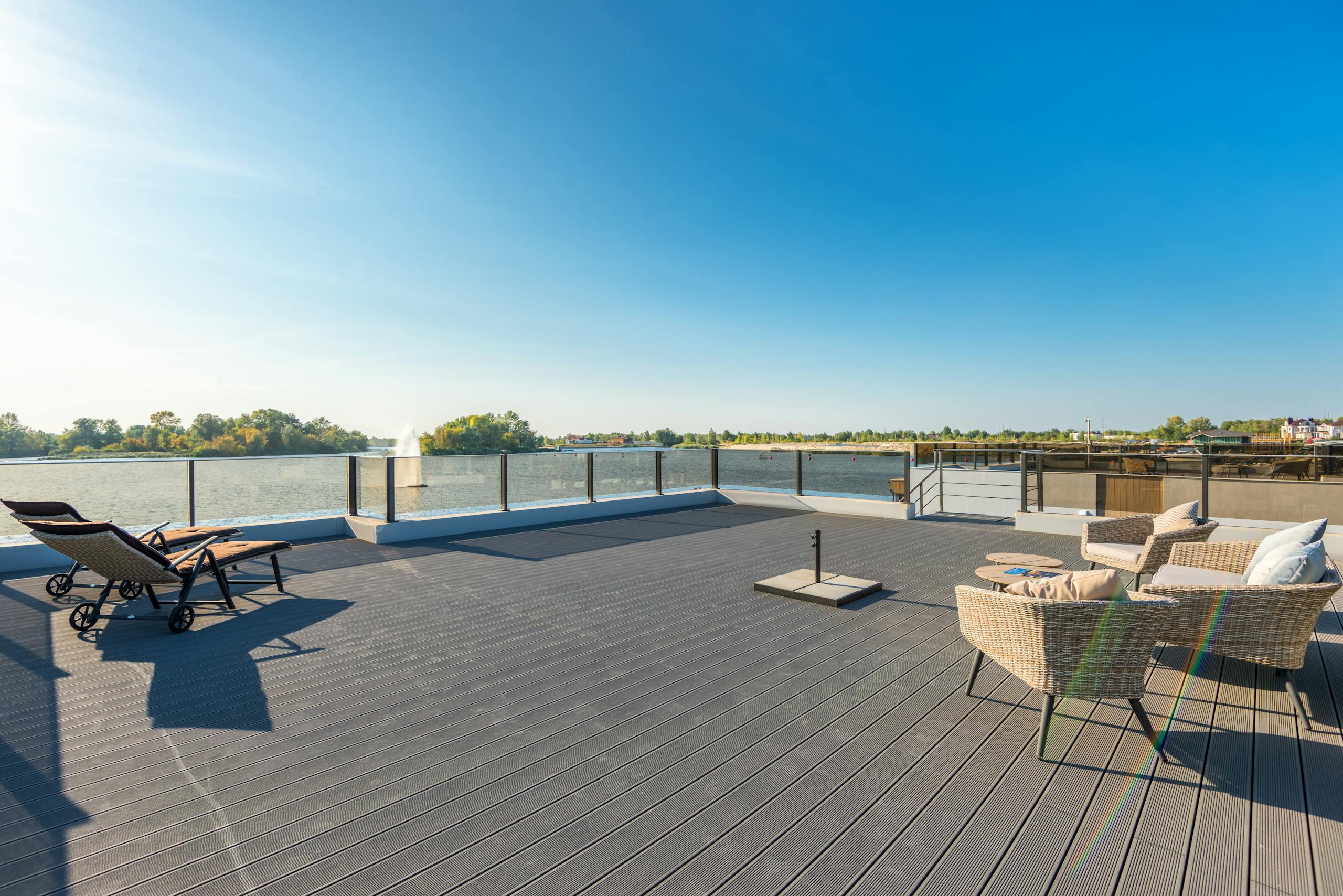 Spacious, sunny waterfront deck with modern wicker seating and lounge chairs overlooking a serene lake.